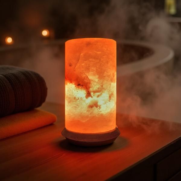 Cylindrical Himalayan salt lamp glowing amber on a wooden spa table with steam and candles in the background