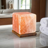 Cube-shaped Himalayan salt lamp on a wooden base on a marble bathroom counter beside rolled towels