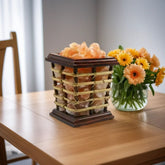 Square wood-and-cane salt lamp basket glowing amber on a dining table beside a vase of orange gerbera daisies