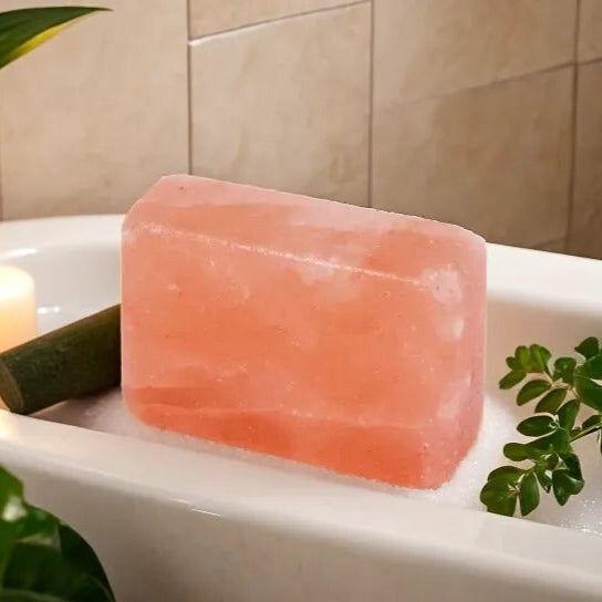 Rectangular Himalayan salt soap bar resting on the edge of a bathtub beside a candle and greenery