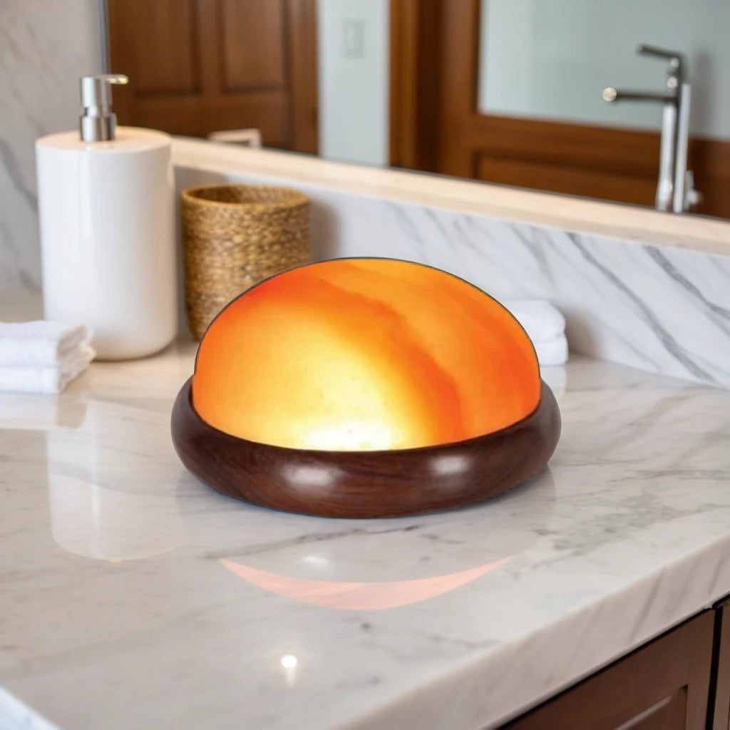 Dome-shaped Himalayan salt massage stone glowing amber in a wooden bowl on a marble bathroom counter